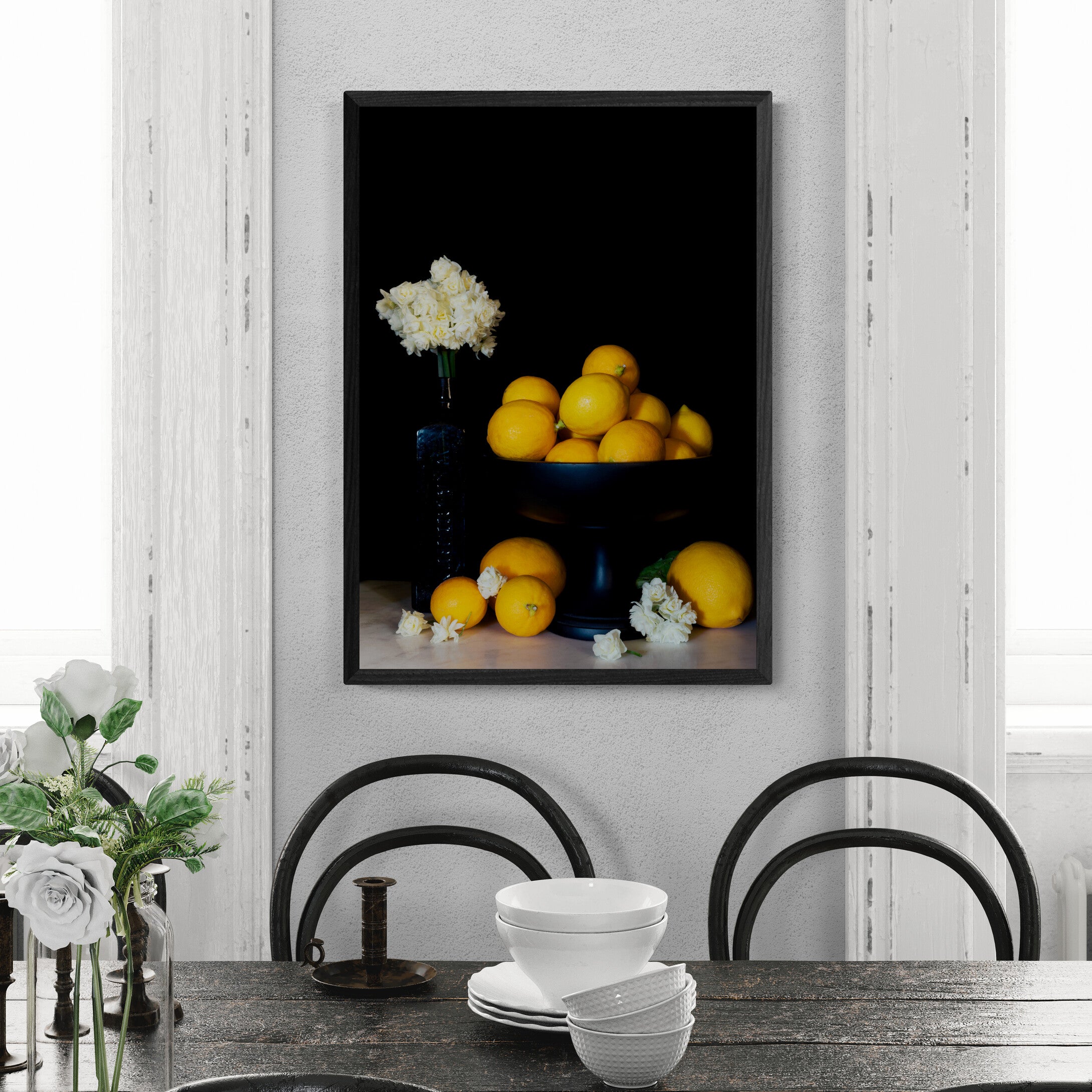 A framed vertical still life art print depicting yellow lemons on a dark pedestal with white flowers, hung on a light gray wall above a dining table with chairs and white dishes.